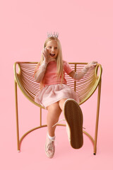 Cute little girl with tiara sitting in armchair on pink background