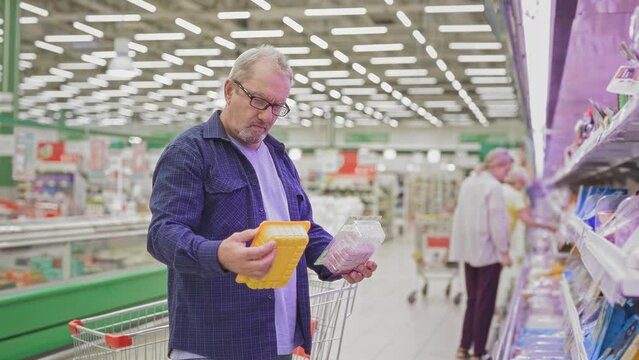 An elderly man standing near the shelves in a supermarket holding meat in plastic packaging, looking at the shelves, comparing prices. Pensioner in a grocery shop