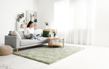 Beautiful young Asian woman with laptop sitting on sofa in living room