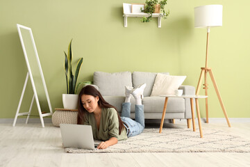 Beautiful young Asian woman with laptop lying on floor in living room
