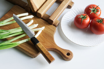 Sharp knife, green onions, vintage solid oak wooden cutting boards and fresh tomatoes in a plate on a light background. Preparing a rustic summer vegetable salad. Photo. Selective focus.