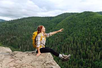 Beautiful traveler with a yellow hiking backpack observes the mountain scenery from a cliff, a view of the European troll tongue. Concept of nature, adventure, freedom.