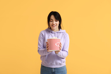 Happy young woman with bucket of popcorn on yellow background