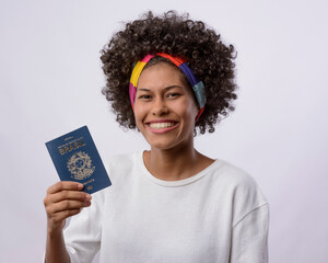 Brazilian passport. Young black woman with black power curly hair, white t-shirt, smiling and holding a Brazilian passport.