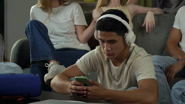 Video of a boy, guy, sitting in a group of teens, young people, listening to music in his headphones, then sharing a song he found with friends through a speaker.