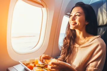 Poster Beautiful young woman eating a plane meal at a window seat, enjoying a meal on a plane ride © VisualProduction