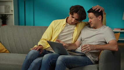 Shot of a homosexual couple at home. One is sitting on the couch, working on the laptop, explaining something to his partner that sits next to him. They are hugging.