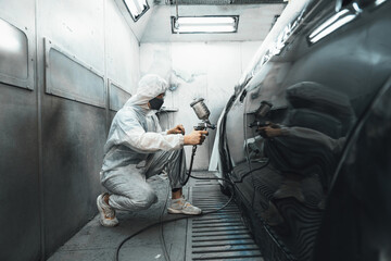 Automotive service worker in full protective gear expertly apply color paint in to car's bodywork with spray gun or respirator painting in chamber workshop. Car paint service for scratch refinish.Oxus