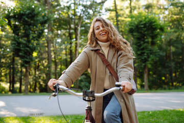 Fototapeta na wymiar Young smiling woman in casual stylish clothes rides a bicycle in a sunny park. Active urban healthy lifestyle concept.