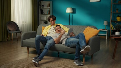 Obraz premium Full-size shot of a homosexual couple at home. They are laying on the couch, watching photos or videos on a tablet, hugging, smiling. One is listening to music.