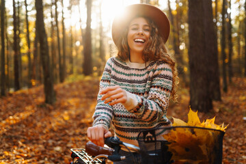A beautiful woman in stylish clothes and a hat walks and rides a bike in a sunny autumn park....