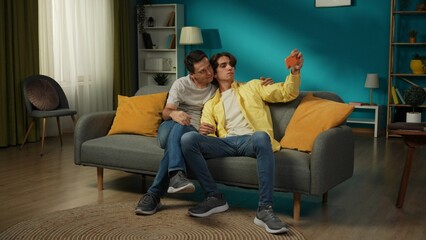 Full-size shot of two homosexual men, couple at home. They are sitting on the couch, hugging, taking selfies, expressing love and affection to each other.