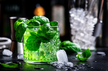 Basil smash green alcoholic cocktail drink with dry gin, sugar syrup, lemon, basil and ice, dark bar counter background