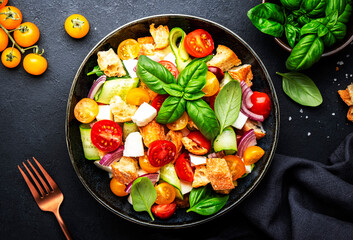 Panzanella salad with stale ciabatta bread, colorful tomatoes, soft cheese, red onion, olive oil, sea salt and green basil, black table background, top view