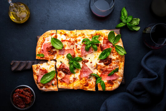 Hot homemade roman pizza with ham, mozzarella cheese, sun dried tomatoes, sauce and green basil, black table background, top view