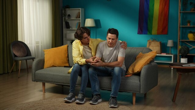 Full-size shot of a homosexual, pansexual, bisexual male couple sitting on the couch, hugging, holding hands. One is comforting their partner. - Powered by Adobe