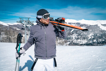 Young male skier walking on top of mountain while holding skies. Handsome man wearing ski suit...