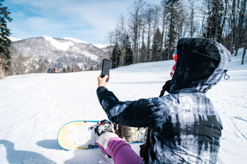Unrecognizable female snowboarder sitting on ground and taking a break while using mobile phone for...