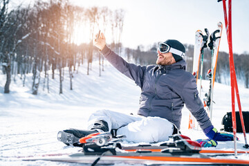 Male skier sitting on snow taking a break while using phone. Smiling man wearing cap and ski...