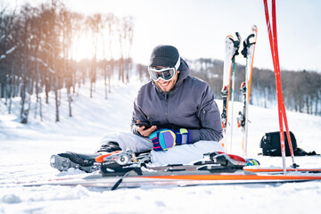 Fototapeta na wymiar Smiling male skier sitting on snow taking a break while using phone and texting. Happy athletic man scrolling his cellphone and feeling satisfied.