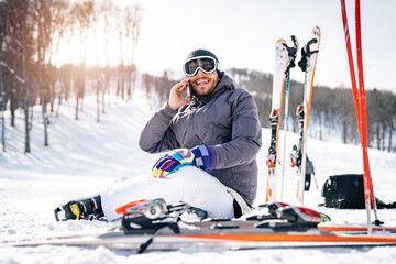 Smiling confident male skier sitting on snow taking a break while using cell phone and having...
