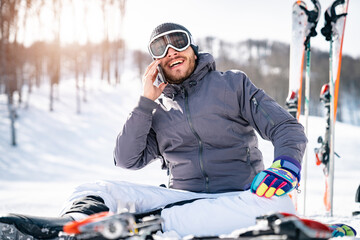 Smiling male skier sitting on snow taking a break while talking on mobile phone. Cheerful...