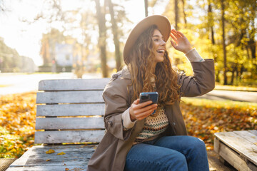 Cute lady in a hat and coat holds a smartphone in her hands while sitting on a bench in an autumn park. Concept of vacation, technology, weekend.