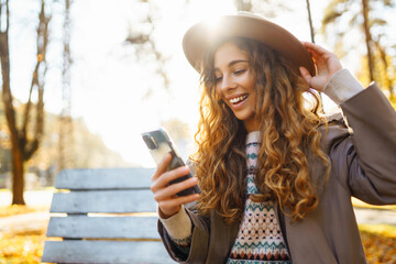 Cute lady in a hat and coat holds a smartphone in her hands while sitting on a bench in an autumn park. Concept of vacation, technology, weekend.
