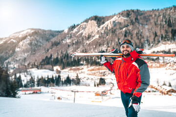 Young confident smiling male skier walking uphill while holding skies. Beautiful winter scenery...