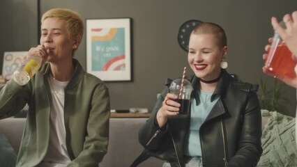 Medium shot of group of multiethnic young people having fun toasting with beer bottles and plastic cocktail cups, getting together in bright modern apartment at daytime