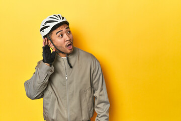 Asian man in bike helmet and gloves, yellow studio backdrop trying to listening a gossip.