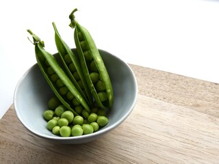 Garden peas (Pisum sativum) and three pea pods in a blue ceramic bowl, on a wooden chopping board. Isolated on a white background. Landscape orientation.