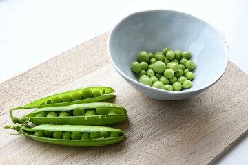 Garden peas (Pisum sativum) in a blue ceramic bowl with pea pods, on a wooden chopping board. Isolated on a white background. Landscape orientation.