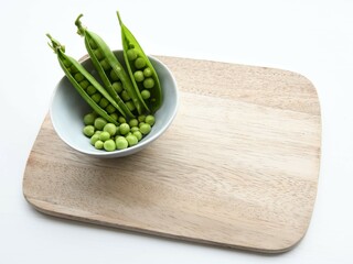 Garden peas (Pisum sativum) and three pea pods in a blue ceramic bowl, on a wooden chopping board. Isolated on a white background. Landscape orientation.