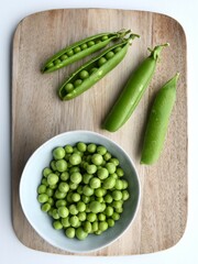 Fresh ingredients on a wooden chopping board with a white background. Garden peas (Pisum sativum) in the pod and in a bowl, taken from above, in portrait orientation.