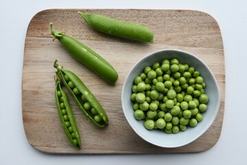 Fresh ingredients on a wooden chopping board with a white background. Garden peas (Pisum sativum) in the pod and in a bowl, taken from above, in landscape orientation.