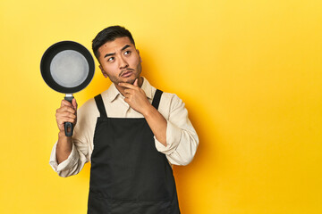 Asian chef holding a pan, yellow studio backdrop looking sideways with doubtful and skeptical expression.