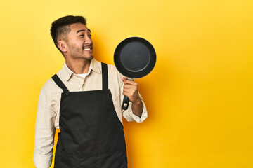 Asian chef holding a pan, yellow studio backdrop looks aside smiling, cheerful and pleasant.