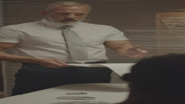 Vertical medium shot of mature gray haired Caucasian police officer interviewing African American suspect man sitting at table with handcuffed hands in interrogation room