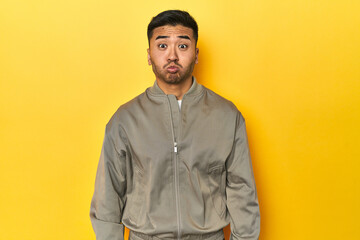 Stylish Asian man in gray jacket on yellow studio blows cheeks, has tired expression. Facial expression concept.