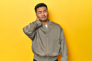 Stylish Asian man in gray jacket on yellow studio having a neck pain due to stress, massaging and touching it with hand.