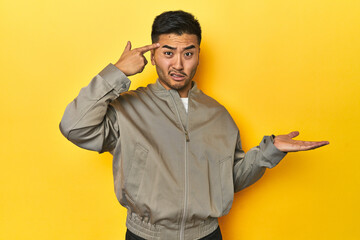 Stylish Asian man in gray jacket on yellow studio holding and showing a product on hand.