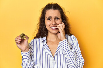 Woman holding a physical Bitcoin coin, biting fingernails, nervous and very anxious.