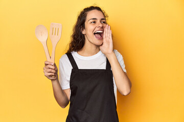 Woman with apron, wooden cooking utensils, yellow, shouting and holding palm near opened mouth.