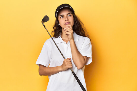 Golfer woman with cap, golf polo, yellow studio, looking sideways with doubtful and skeptical expression.