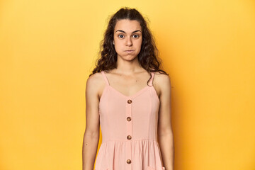 Young Caucasian woman, yellow studio background, blows cheeks, has tired expression. Facial expression concept. - obrazy, fototapety, plakaty