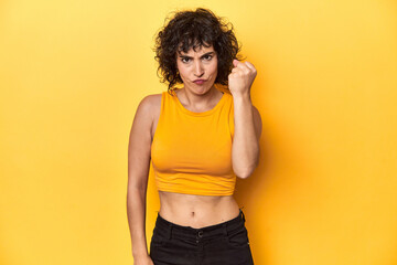 Curly-haired Caucasian woman in yellow top showing fist to camera, aggressive facial expression.