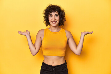 Curly-haired Caucasian woman in yellow top makes scale with arms, feels happy and confident.