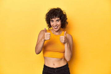 Curly-haired Caucasian woman in yellow top raising both thumbs up, smiling and confident.