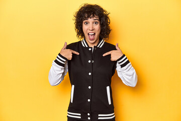 Curly-haired Caucasian woman in baseball jacket surprised pointing with finger, smiling broadly.